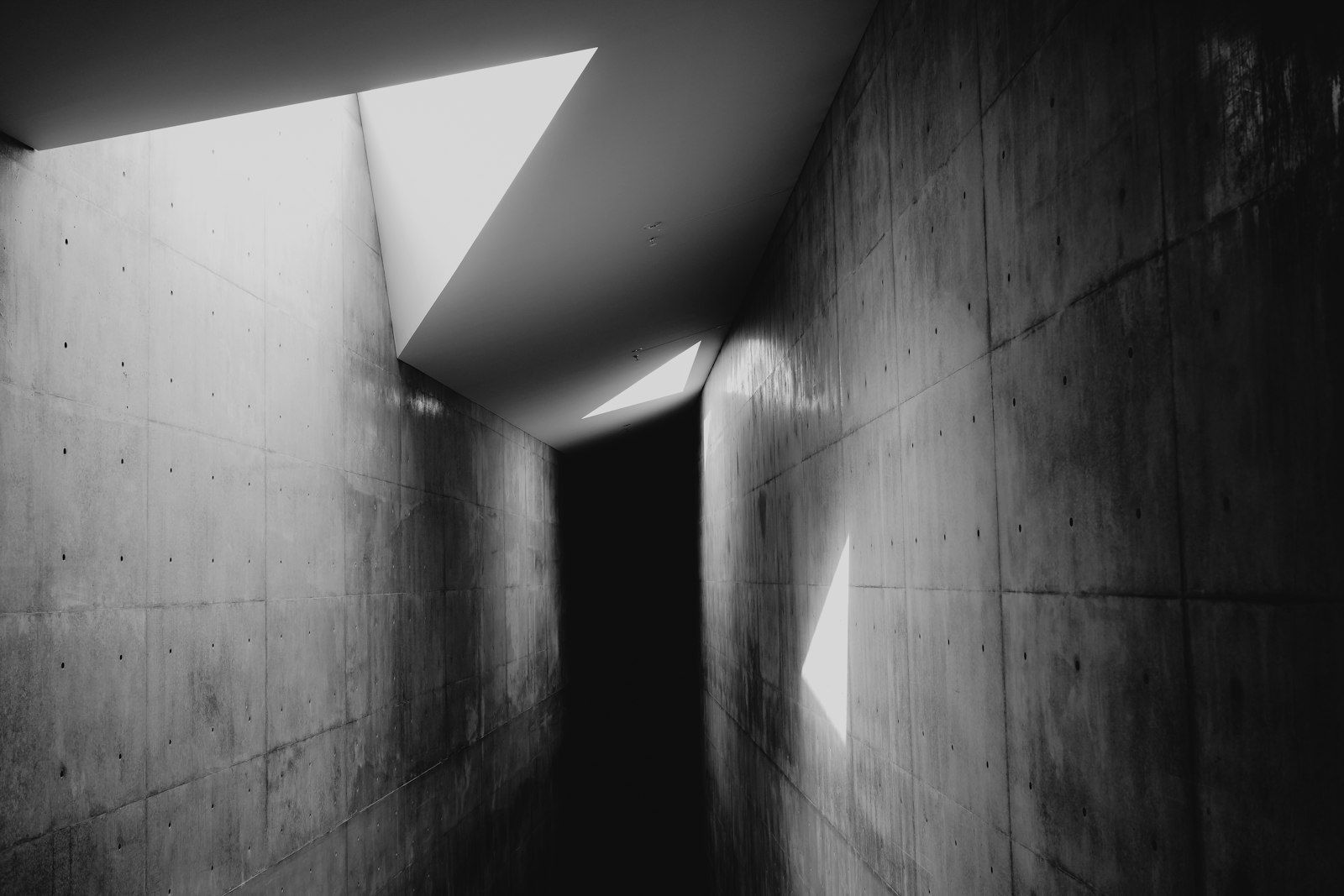 Skylight cutting hard bands of light and shadow down a bare concrete hallway.